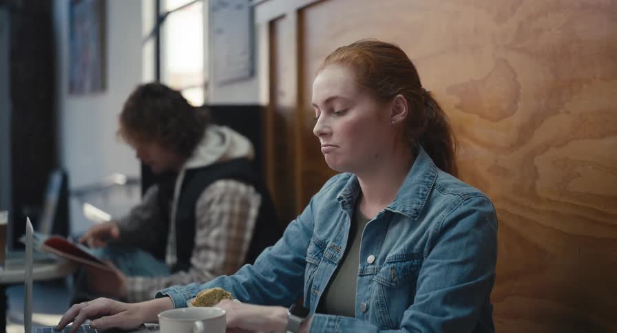 A focused young woman wearing earbuds works on her laptop and enjoys a snack in a modern cafe