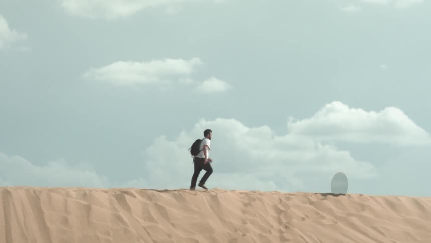 A man hikes over a sand dune towards a mirror