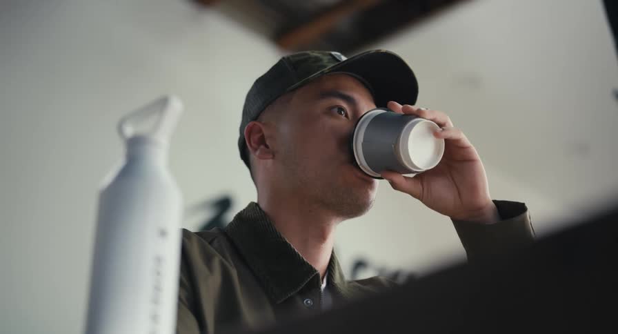 A young man in a cap drinks from a coffee cup and talks while sitting at his desk in a modern workspace