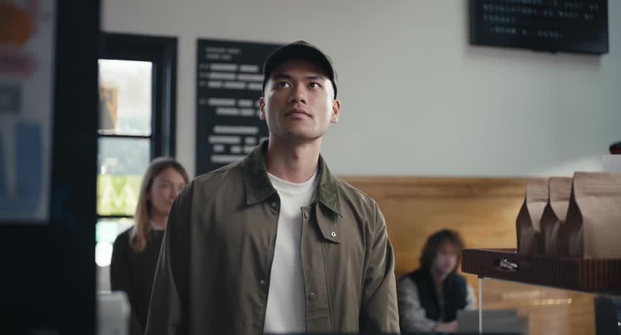 A young man in a baseball cap walks up to the counter in a modern coffee shop, places his order, and receives his to-go coffee