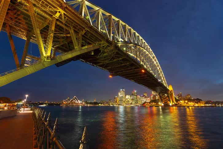 A dramatic day-to-night timelapse from beneath the Sydney Harbour Bridge captures the iconic city skyline and bustling harbor traffic as the vibrant city lights illuminate the night