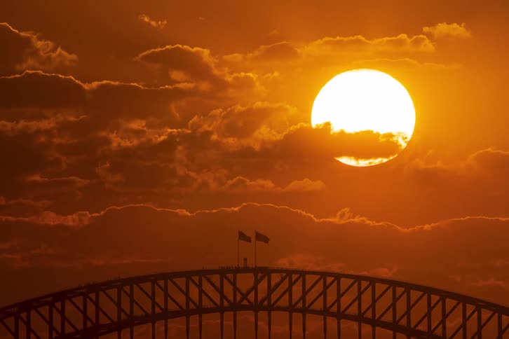 A stunning time-lapse captures the large, fiery orange sun setting behind dramatic clouds over the iconic Sydney Harbour Bridge silhouette