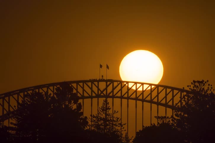 A dramatic timelapse shows the sun setting behind the iconic Sydney Harbour Bridge against a hazy orange sky