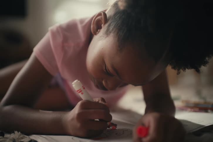A focused young girl lies on the floor of a sunlit room while coloring in a book with a red marker