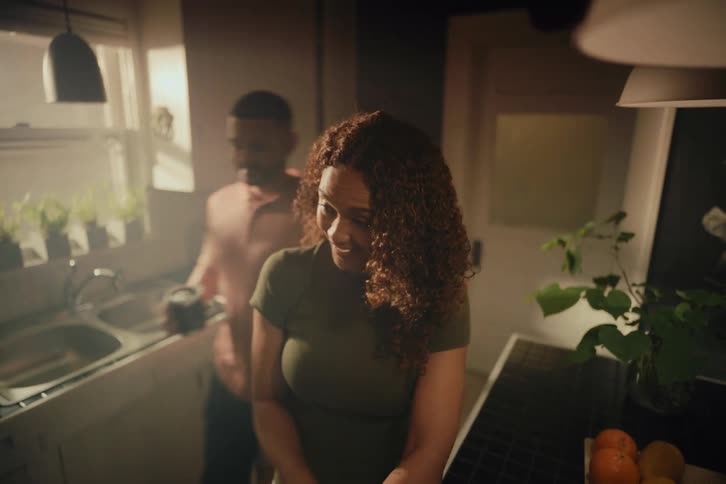 A caring man surprises his partner from behind with a gentle touch, making her laugh in their cozy, sun-kissed kitchen