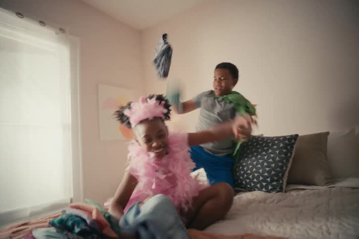 Happy siblings in costume play dress-up and throw clothes everywhere on a bed in slow motion