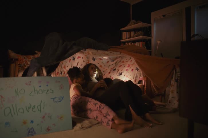 A mother and her two children snuggle together in a cozy blanket fort at night, lit by the glow of a phone
