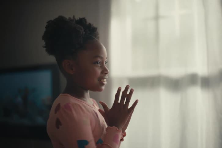An excited young girl in pink pajamas claps her hands with joy while looking out a sunlit window