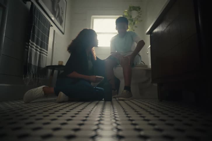 A caring mother tends to her son's scraped knee on the tiled floor of a sunlit bathroom
