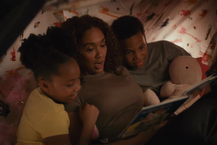 A mother reads a bedtime story to her son and daughter while snuggled together in a cozy blanket fort at night