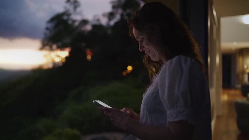 A happy woman smiles while messaging on her smartphone from a balcony overlooking a scenic landscape at dusk