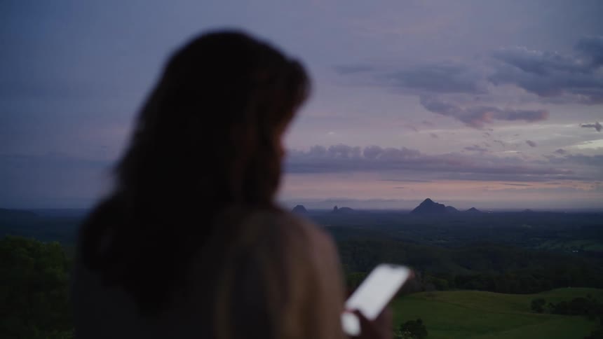 A woman holds her smartphone while enjoying the breathtaking view of a mountain range at twilight