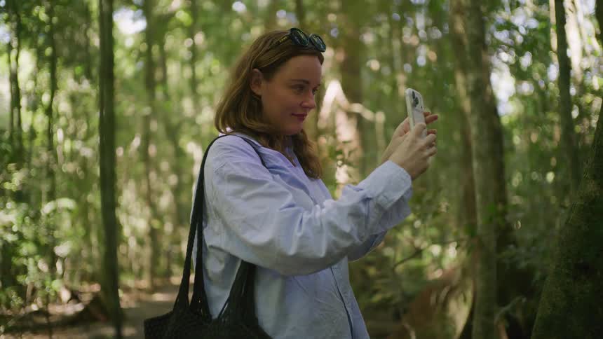 A woman stands in a lush green forest, capturing the beautiful scenery on her smartphone
