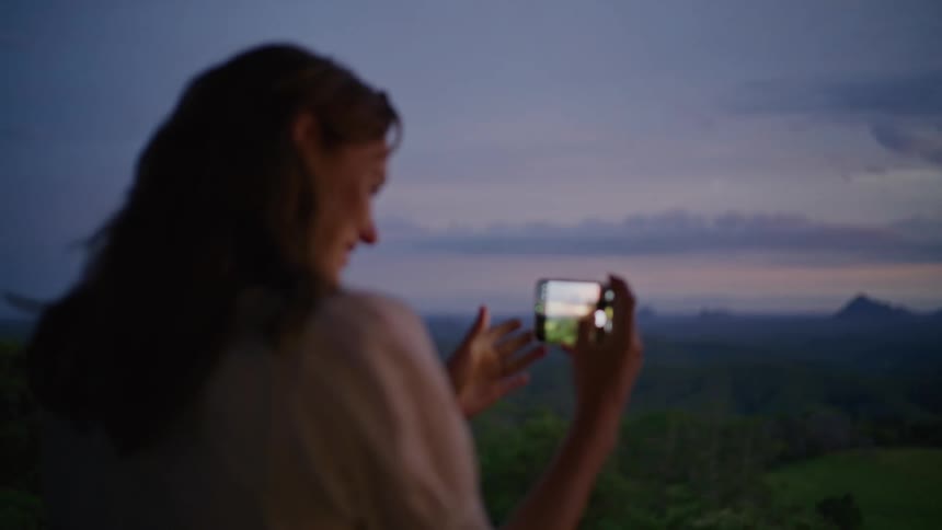 A smiling woman captures a scenic mountain landscape on her smartphone during a beautiful twilight evening
