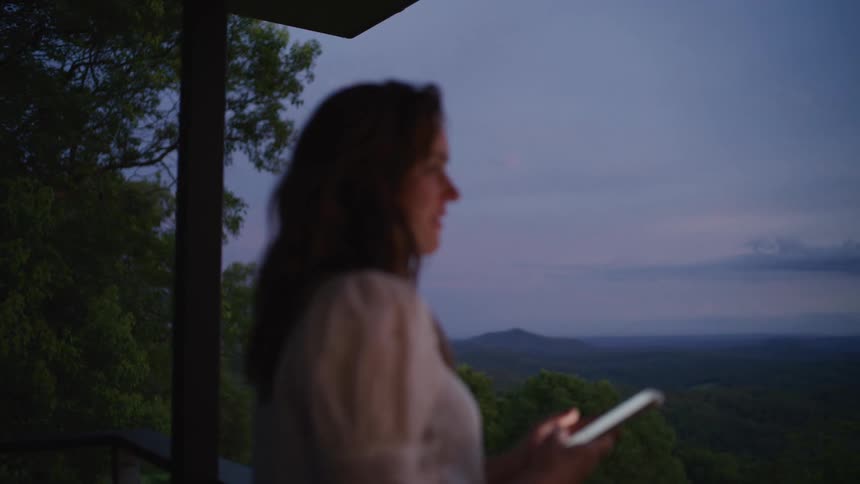 A woman standing on a balcony at dusk uses her smartphone with a beautiful mountain valley in the background