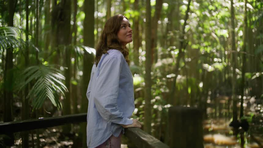 A woman stands on a wooden boardwalk in a lush, sun-dappled rainforest, peacefully admiring the natural beauty around her