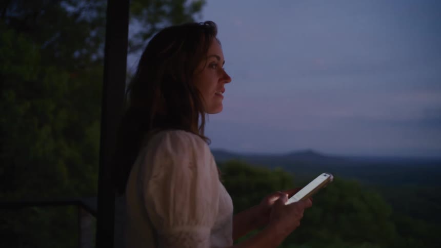 A woman smiles while using her smartphone on a balcony overlooking a beautiful mountain range at dusk