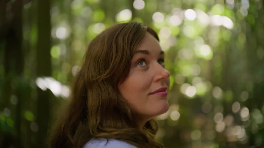 A serene young woman opens her eyes and gazes up in wonder while standing in a lush, green forest