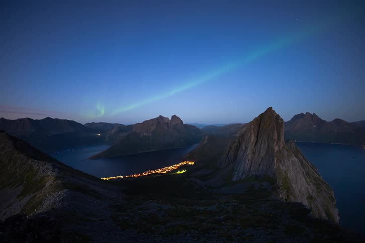A stunning time-lapse captures the Aurora Borealis dancing in the night sky over a rugged mountain range and an illuminated coastal town below