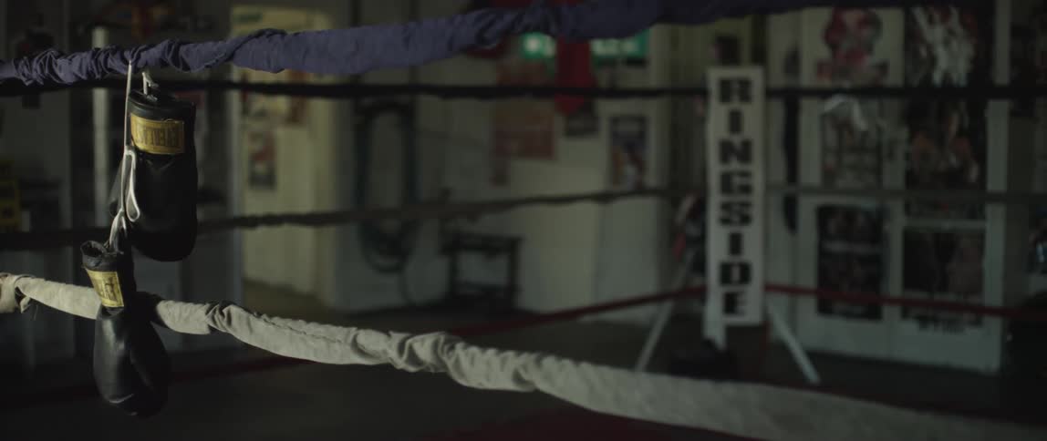 Boxing gloves dangling from the top rope of a boxing ring that has a ringside sign in it