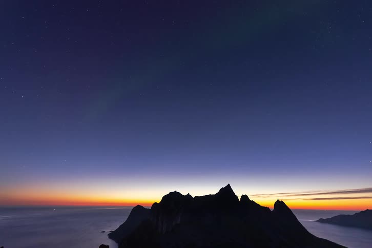 The ethereal green Aurora Borealis dances across a starry twilight sky above a silhouetted mountain range by the ocean