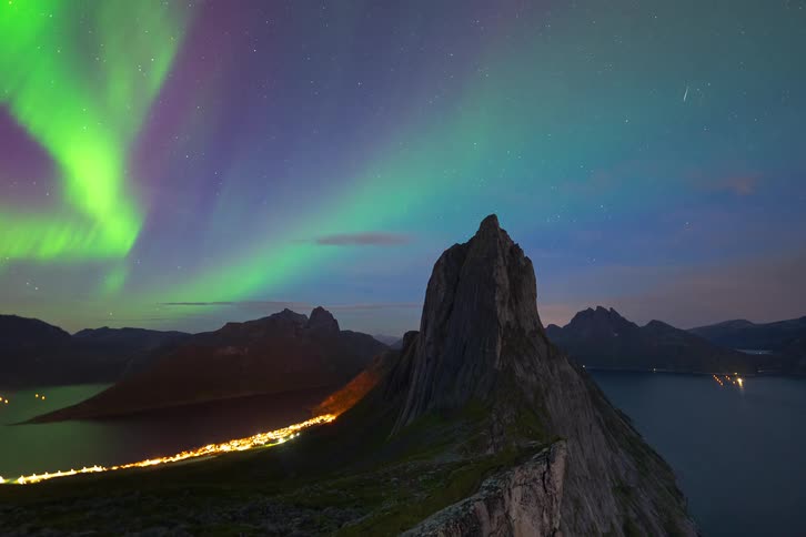 A vibrant timelapse of the green Northern Lights dancing across the starry night sky over a dramatic mountain landscape and a glowing coastal town