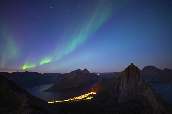A spectacular time-lapse of the vibrant green Aurora Borealis dancing in the starry night sky over a tranquil mountain fjord and a glowing coastal village