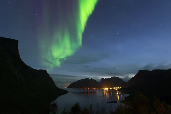 The vibrant green Northern Lights swirl across the starry night sky above a tranquil fjord and a glowing coastal town