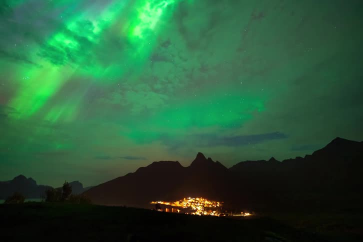 A stunning timelapse of the green and purple Northern Lights dancing in the starry night sky above a glowing village nestled in the mountains