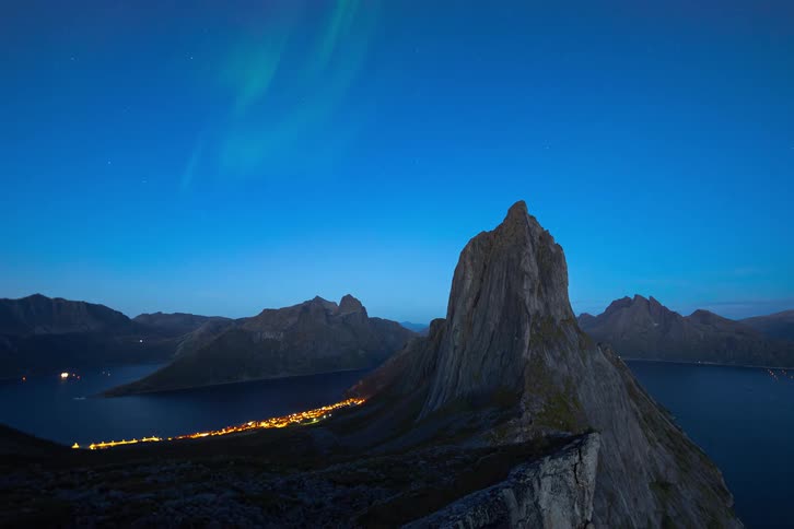 A stunning timelapse captures the vibrant Aurora Borealis dancing in the night sky over a dramatic mountain peak and a glowing coastal town