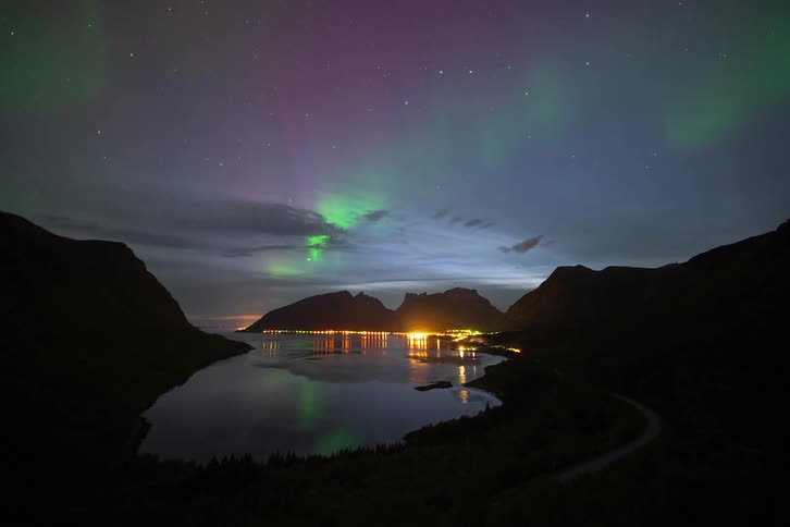A stunning timelapse captures the vibrant green and purple Northern Lights dancing in the starry night sky over a tranquil fjord and an illuminated coastal village