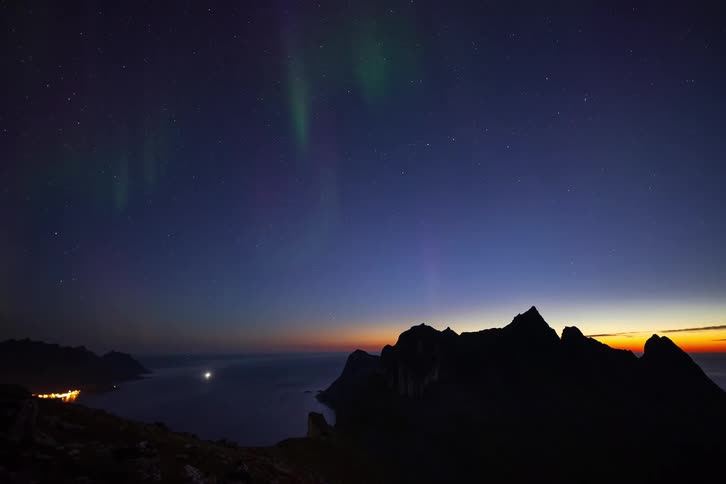 The Aurora Borealis dances across the starry night sky above a silhouetted mountain range overlooking a tranquil fjord at dusk