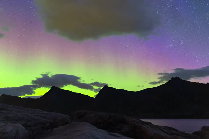 A stunning time-lapse captures the brilliant green and purple Aurora Borealis dancing in the starry night sky over a silhouetted mountain range