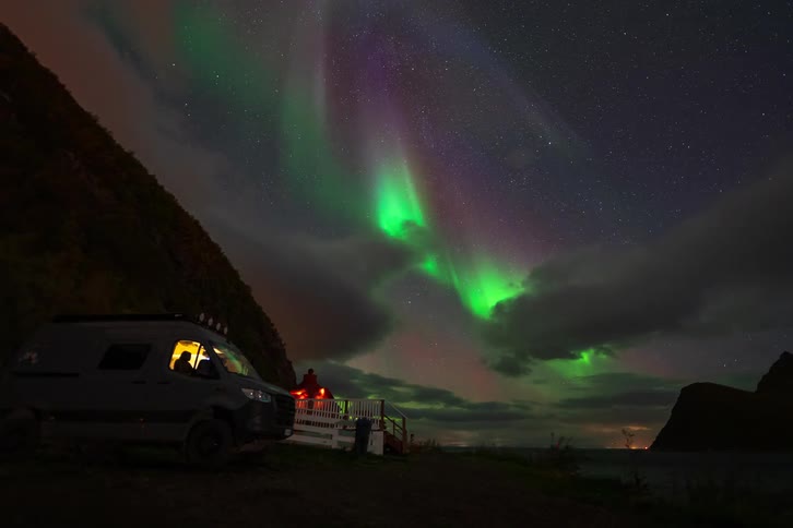 A stunning timelapse shows the vibrant green and purple Aurora Borealis dancing across the starry night sky above a camper van parked by the sea