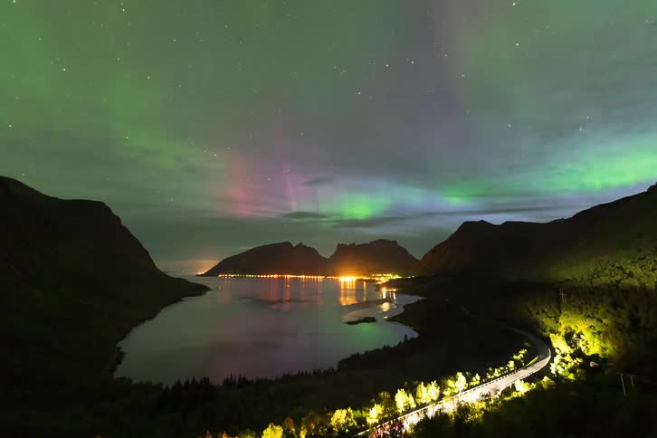 A stunning timelapse of the Aurora Borealis with green and pink lights dancing over a tranquil fjord and a lit coastal town nestled between mountains