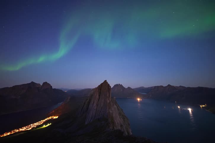A stunning timelapse shows the vibrant green Northern Lights dancing across the starry night sky over a dramatic mountain fjord and an illuminated coastal village