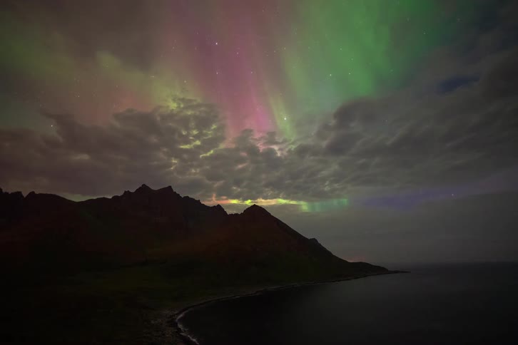 A beautiful time-lapse of the vibrant green and purple Aurora Borealis dancing through the clouds over a dark, mountainous coastline