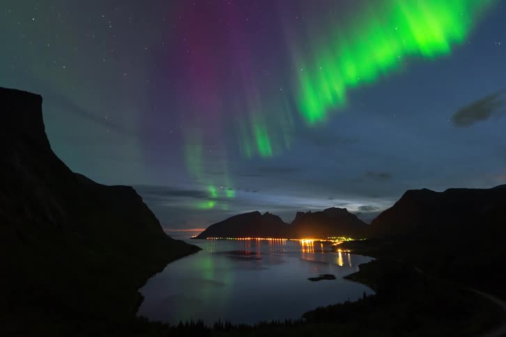 A stunning time-lapse captures the vibrant green and purple Aurora Borealis dancing over a tranquil fjord and an illuminated village nestled between mountains