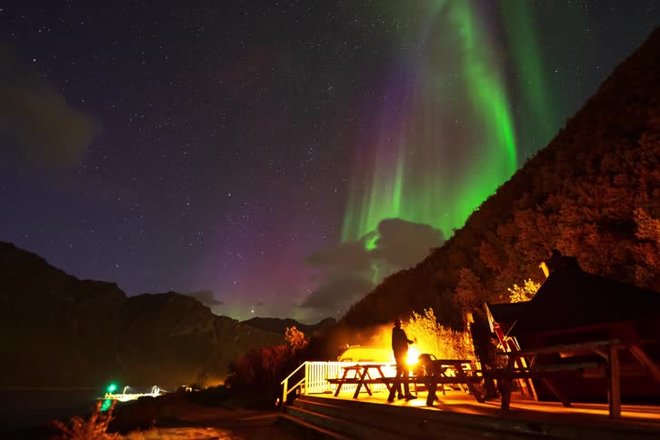 Time-lapse of the vibrant Aurora Borealis dancing in the starry night sky over a mountain cabin where people gather around a bonfire