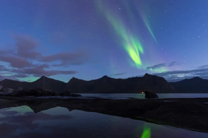 A stunning timelapse shows the vibrant green and purple Aurora Borealis dancing across the starry night sky over a mountain range, reflected in the calm water