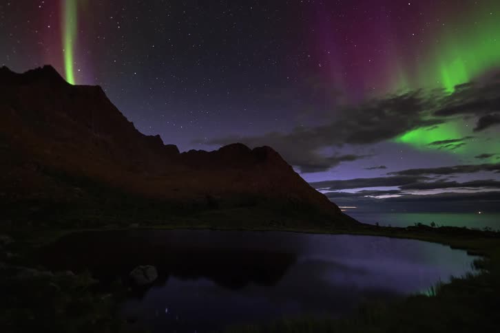 A stunning time-lapse captures the vibrant green and magenta Aurora Borealis dancing in the starry night sky over a tranquil mountain lake