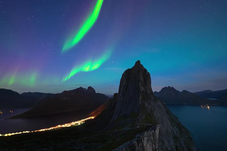 A stunning timelapse of the vibrant green and purple Northern Lights dancing across the starry night sky above a majestic mountain peak and a glowing coastal village