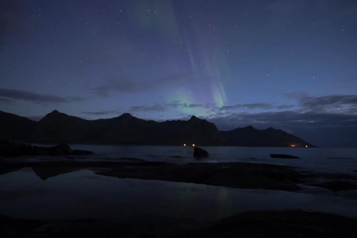 A mesmerizing time-lapse captures the vibrant green and purple hues of the Aurora Borealis dancing across the starry night sky above a silhouetted mountain range and a calm sea