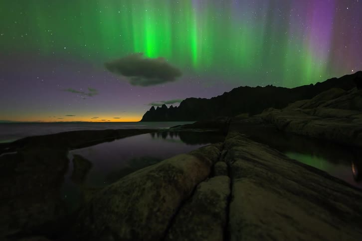 A vibrant timelapse of the Northern Lights dancing across a starry sky over a rocky coast as the last bit of sunset glows on the horizon