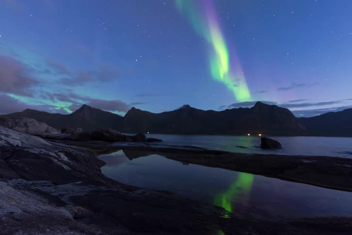A breathtaking timelapse of the green and purple northern lights dancing over a mountain fjord, with its reflection visible in the water below