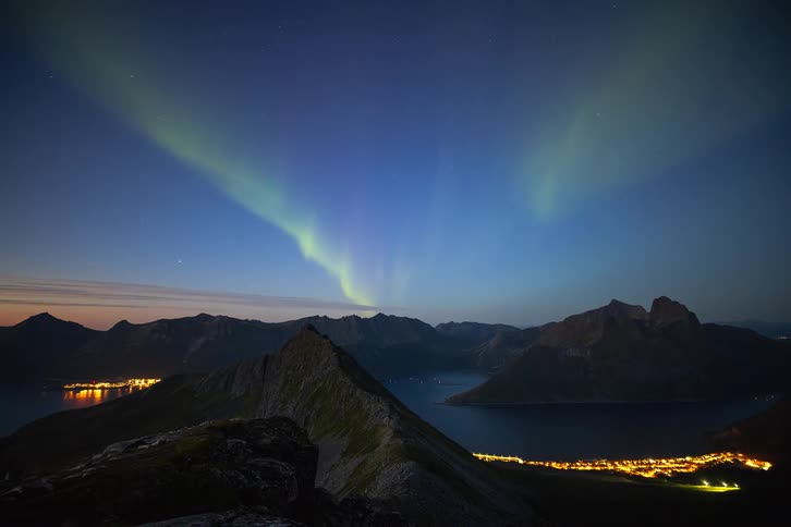 The magical Aurora Borealis dances across the starry night sky above a dramatic mountain landscape and brightly lit coastal villages