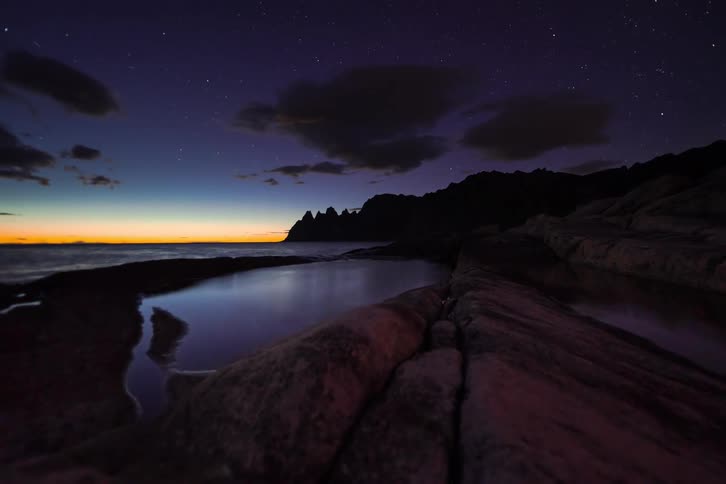 A serene timelapse captures the transition from a colorful twilight to a brilliant starry night over a rocky coastline, with the sky reflected in a calm tidal pool