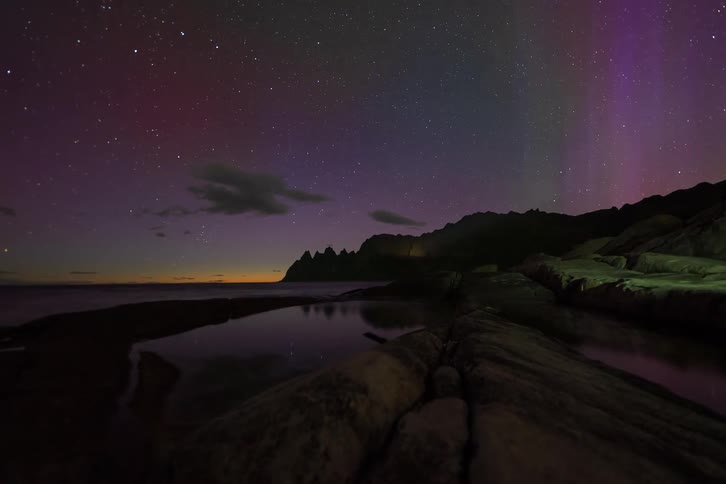 Vibrant green and purple Aurora Borealis dances across the starry night sky over a rocky beach in a stunning time-lapse