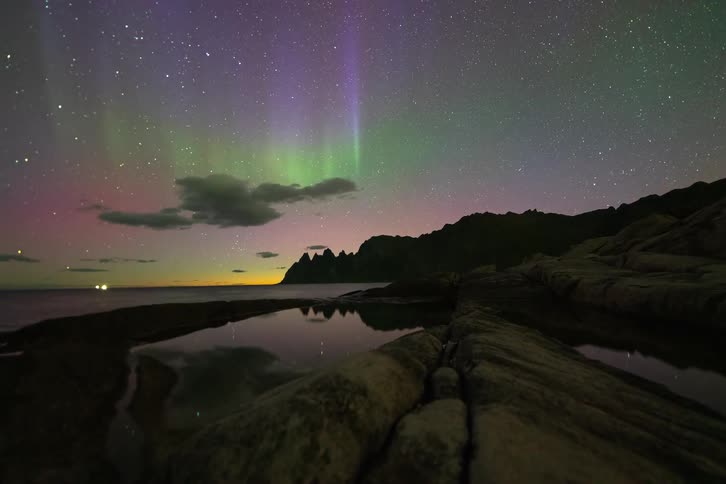 A stunning time-lapse captures the vibrant Aurora Borealis dancing across the starry night sky above a rocky coast with a reflection pool