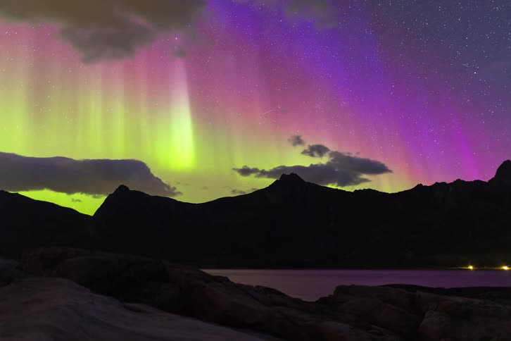 A stunning time-lapse captures the vibrant green and purple Aurora Borealis dancing in the starry night sky over a silhouetted mountain range and a calm lake